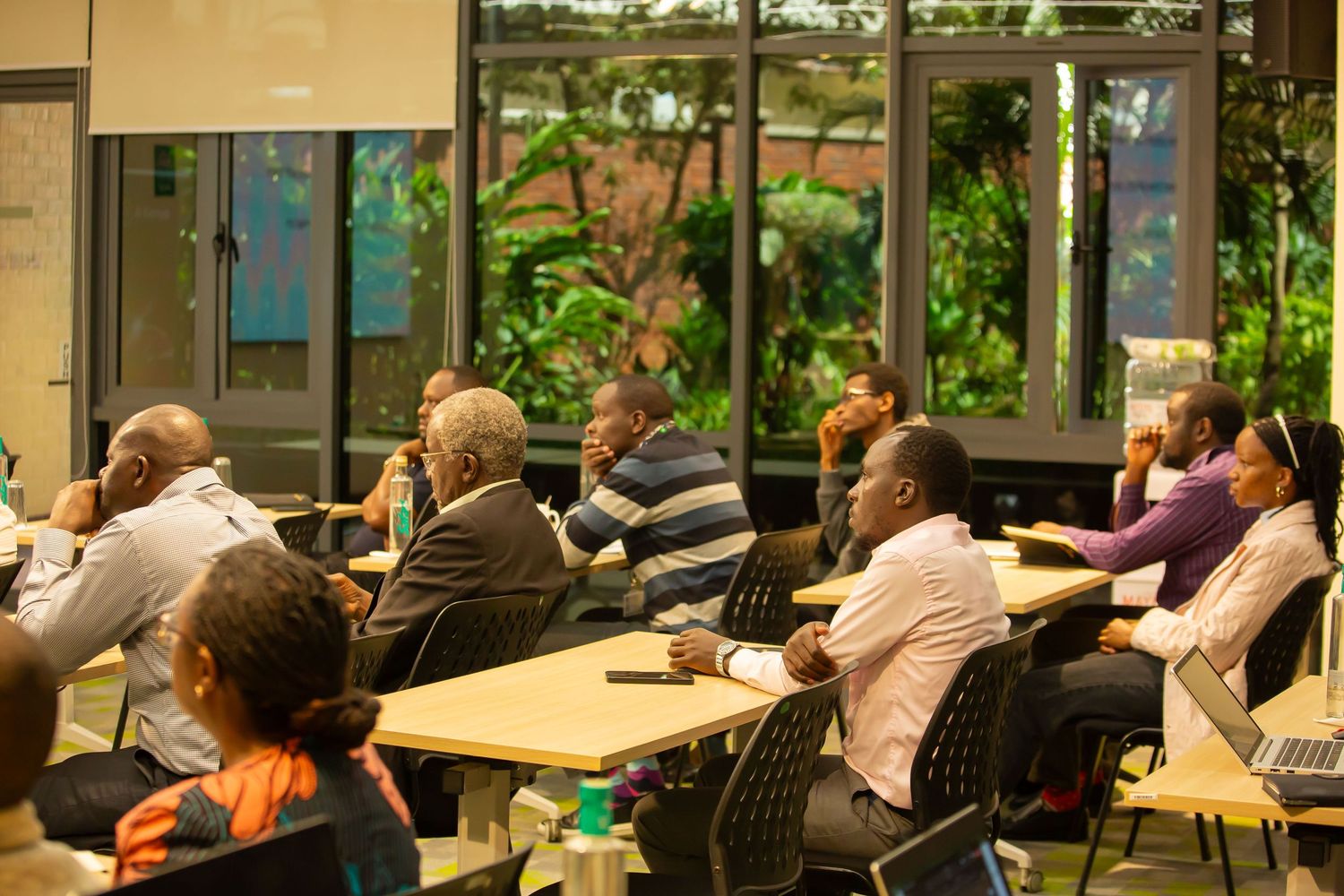 Conference attendees sit in rows of chairs while listening to a presentation in a bright meeting room with glass walls.