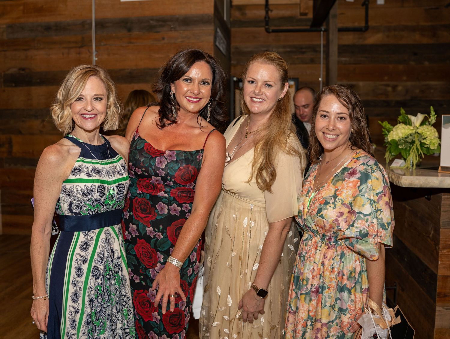 Four people wearing colorful floral and patterned dresses pose together at an event with wooden walls in the background.