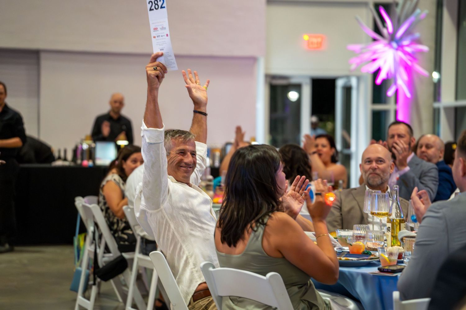 Guests at a lively indoor celebration raise their hands in excitement during a festive dinner event.