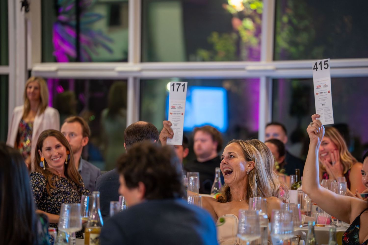 People holding up numbered cards at a social gathering with colorful lighting and table settings.