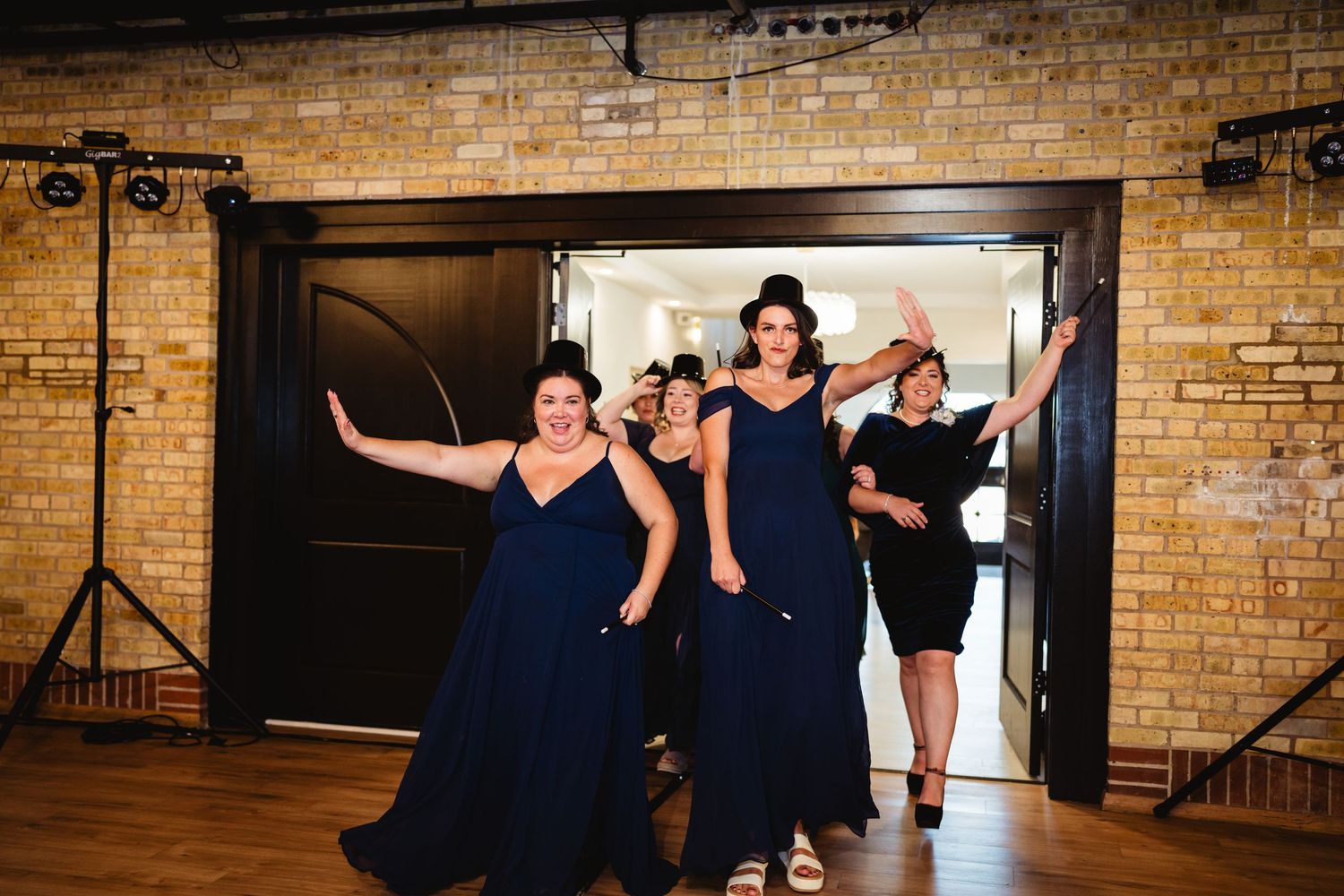 A group in navy blue formal dresses enters through a doorway in a rustic brick-walled venue.