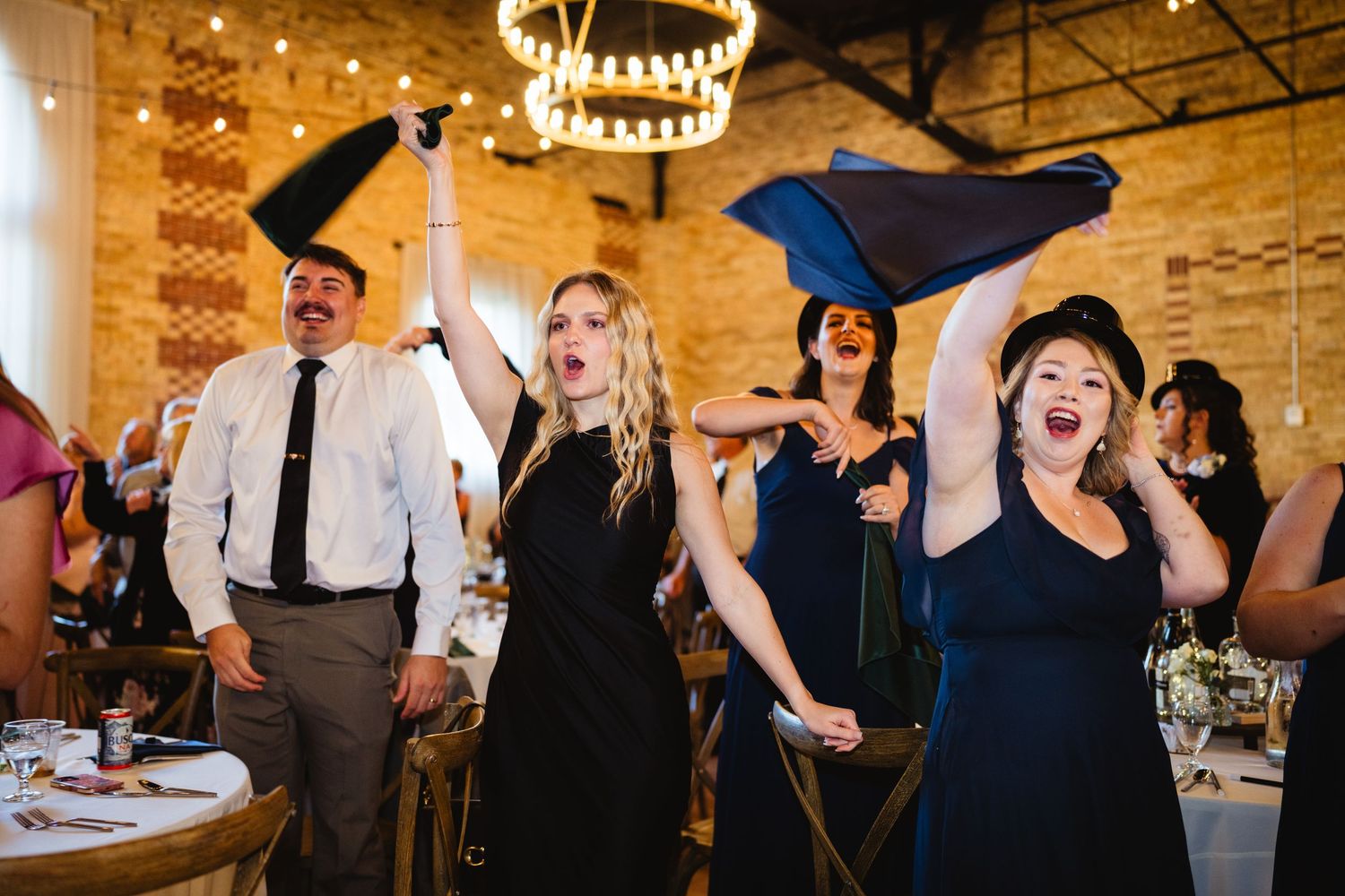 Wedding guests laugh and celebrate during a reception in a rustic venue with wooden beams and warm lighting.
