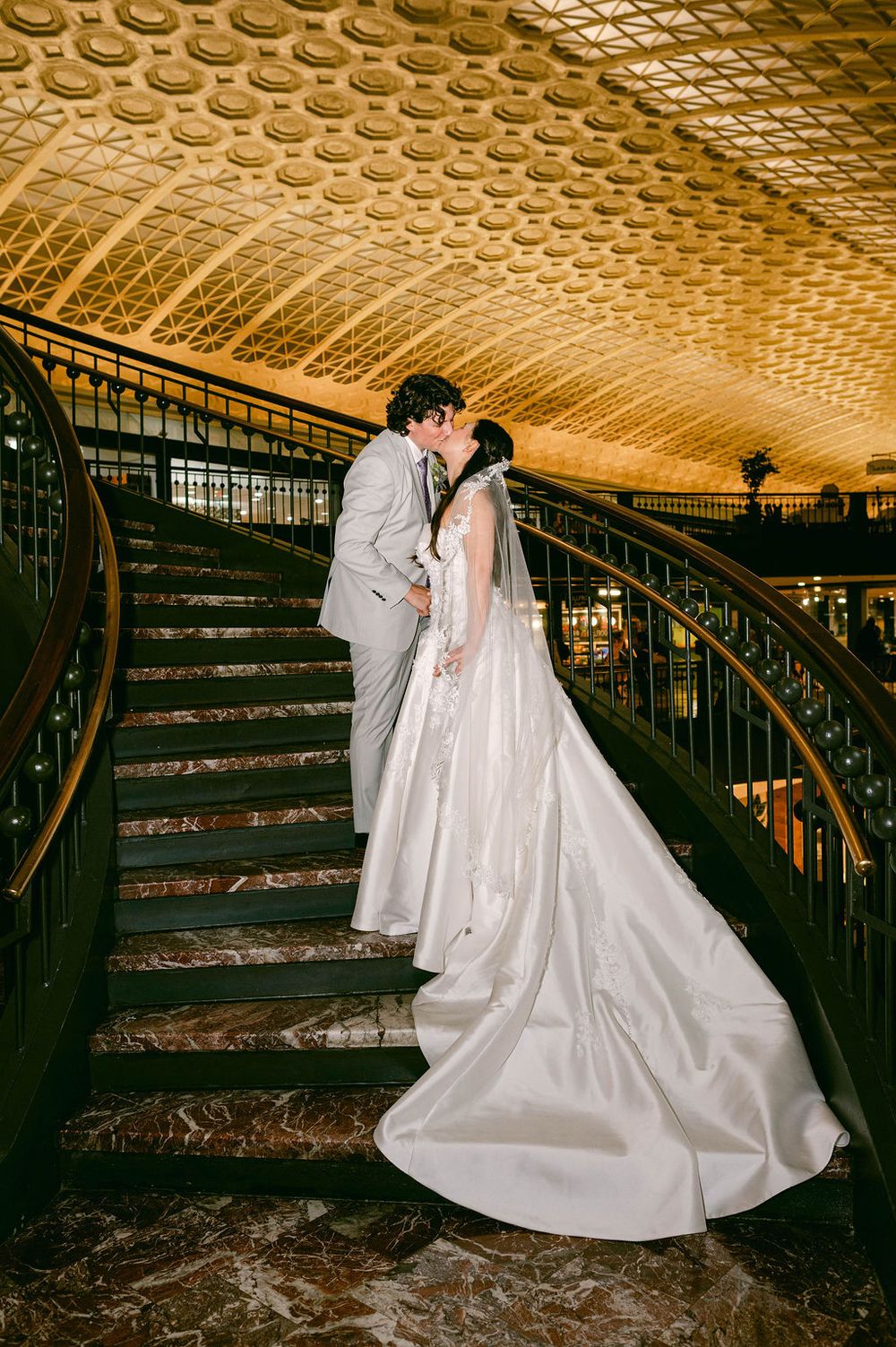 A couple shares an intimate moment on an ornate staircase with a decorative gold ceiling in a historic building.