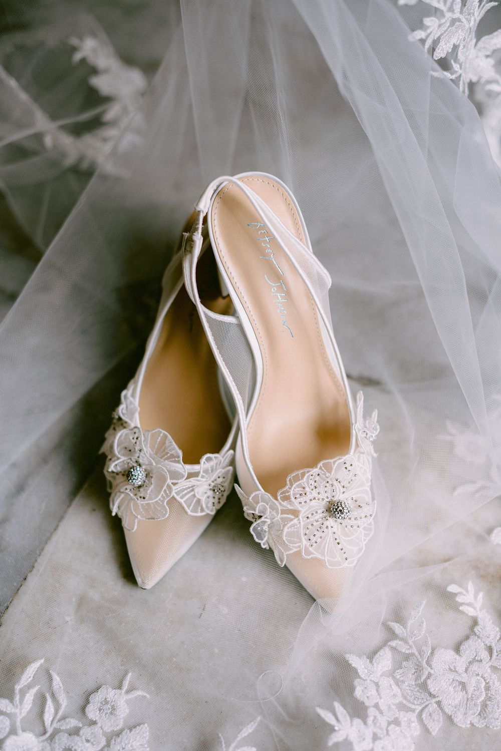 Elegant white lace wedding slingback heels displayed on delicate tulle veil with floral appliques.
