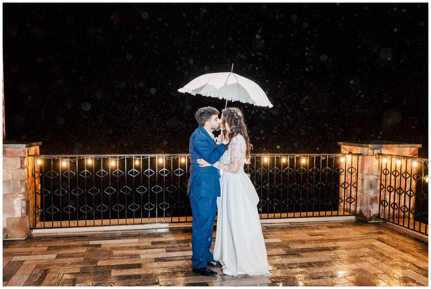 A couple in wedding attire poses with a white umbrella at night on a deck with illuminated railings.