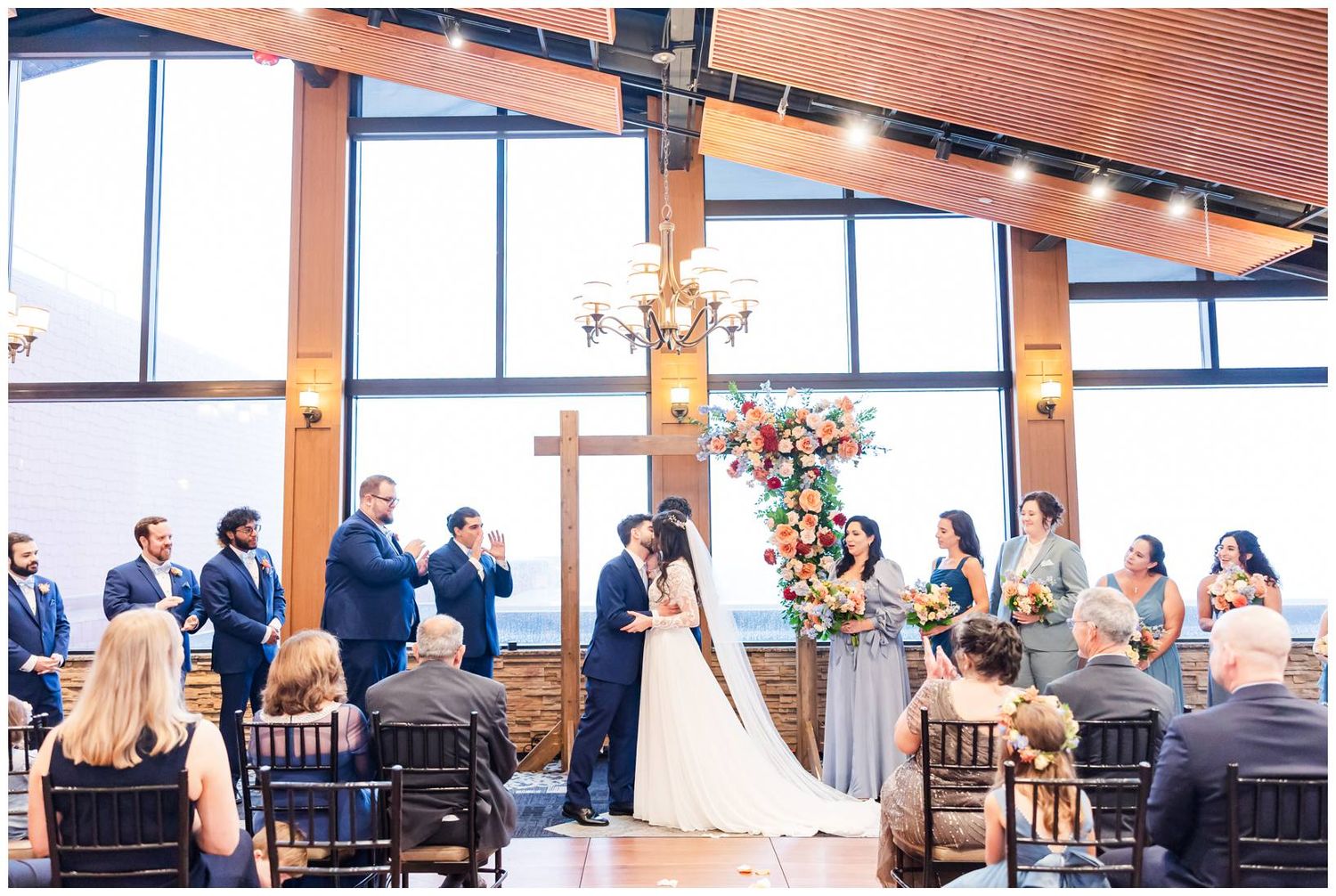 A panoramic view of a rustic wedding ceremony in a wooden beam venue with elegant chandeliers and floral decor.
