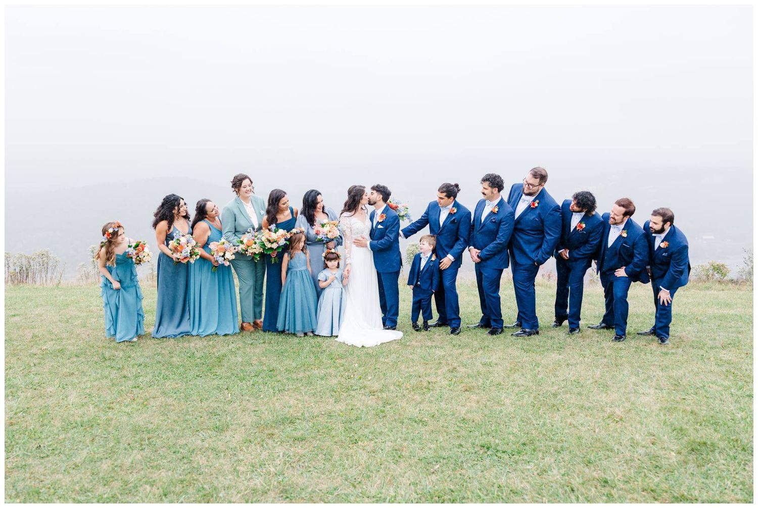 Wedding party in blue attire poses together on a grassy hilltop with mountain views.