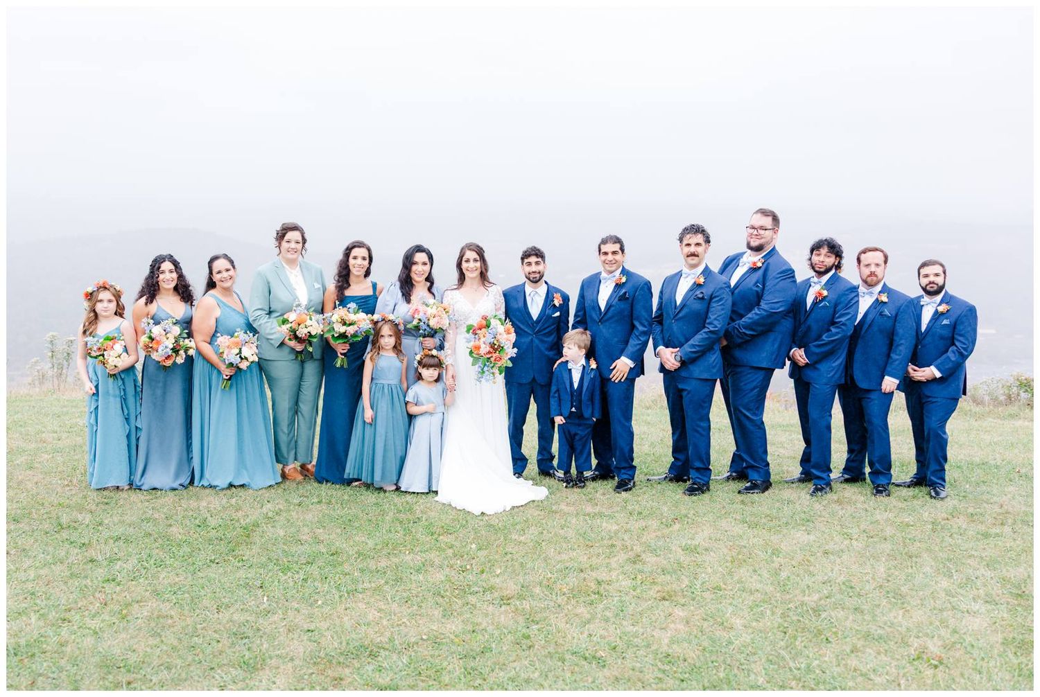 Large wedding party in blue attire posed on grassy hilltop with mountain vista.