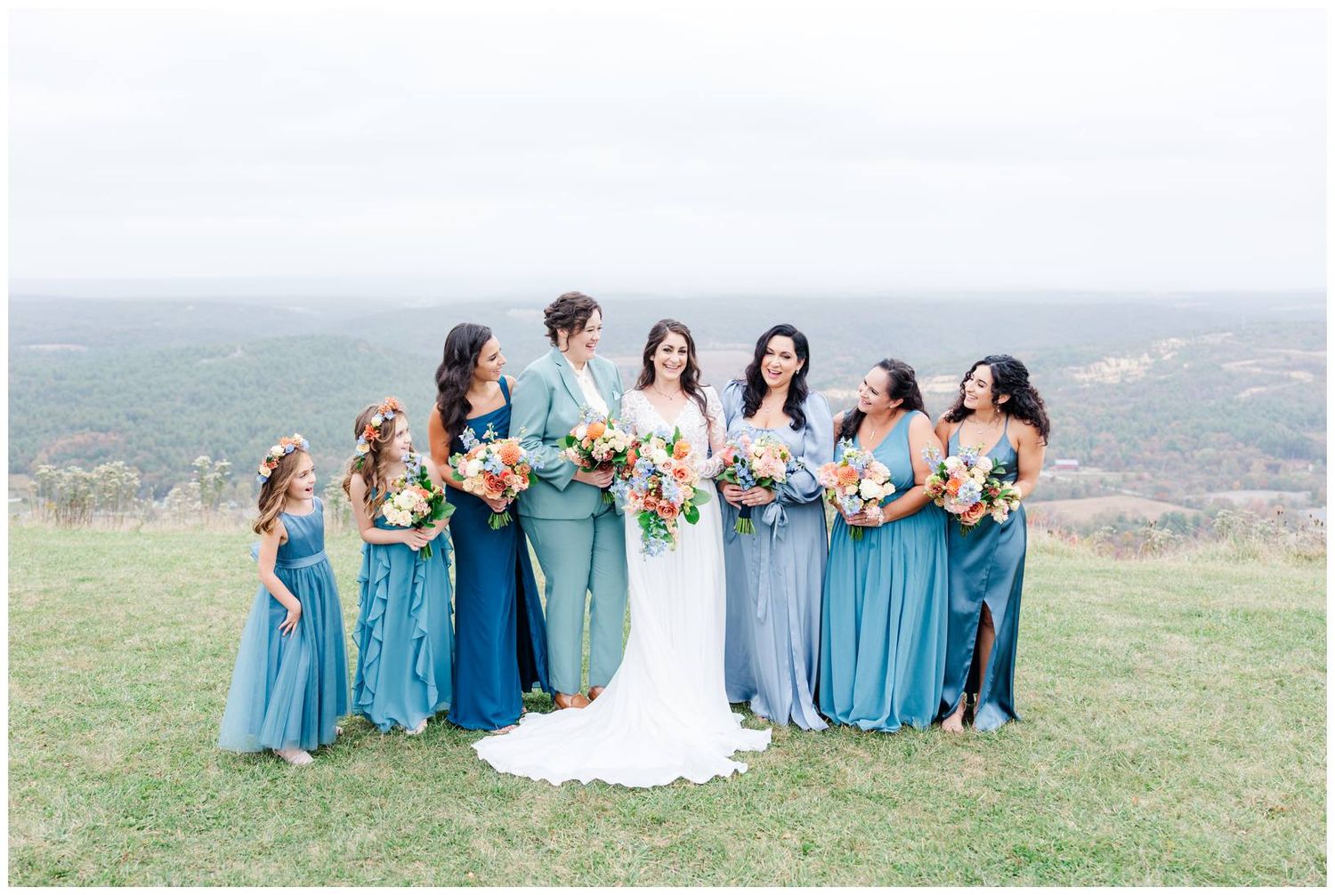 Bridesmaids in flowing blue dresses gather on hilltop for wedding photos with mountain views.