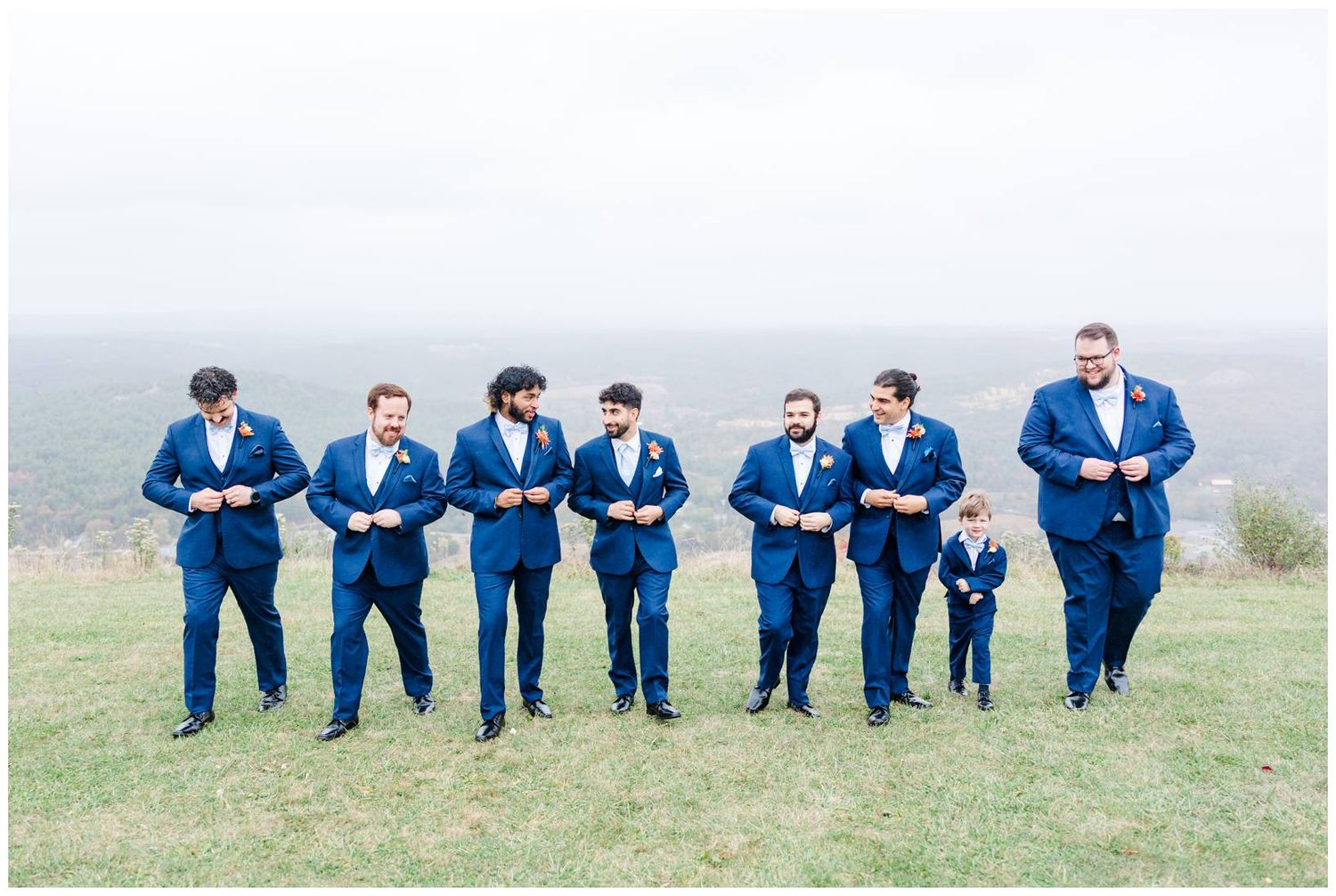 Groomsmen in navy suits lined up on grassy hilltop with mountain backdrop.