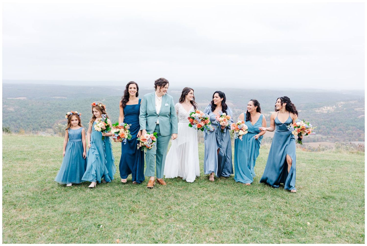 Wedding party in blue dresses and white gown posed on hilltop with mountain views.
