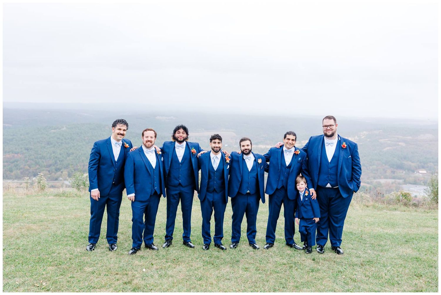 Groomsmen in navy suits pose together on grassy hilltop with scenic mountain backdrop.