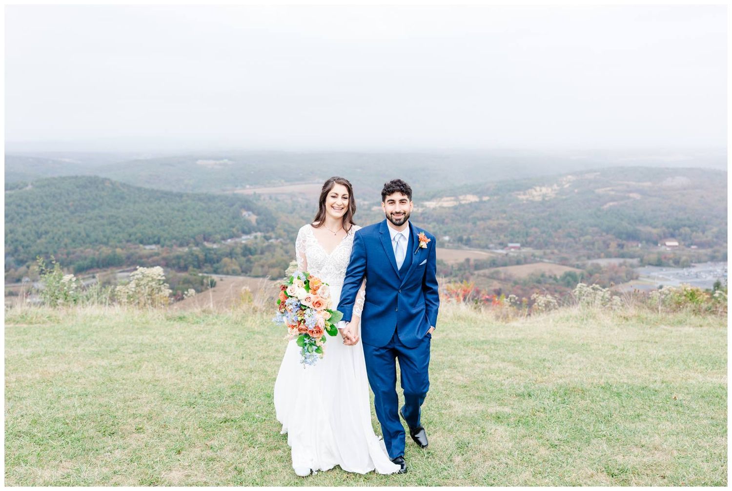 A newlywed couple walks hand in hand across a hilltop meadow with mountain views.