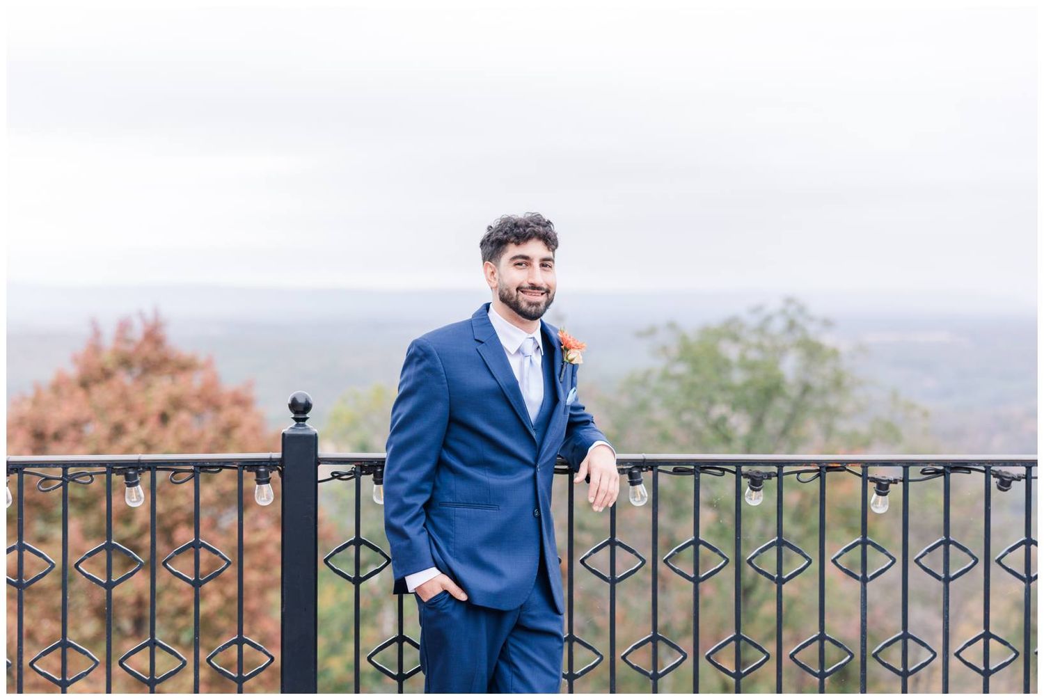 A groom in a navy blue suit poses on a balcony with mountain views in the background.