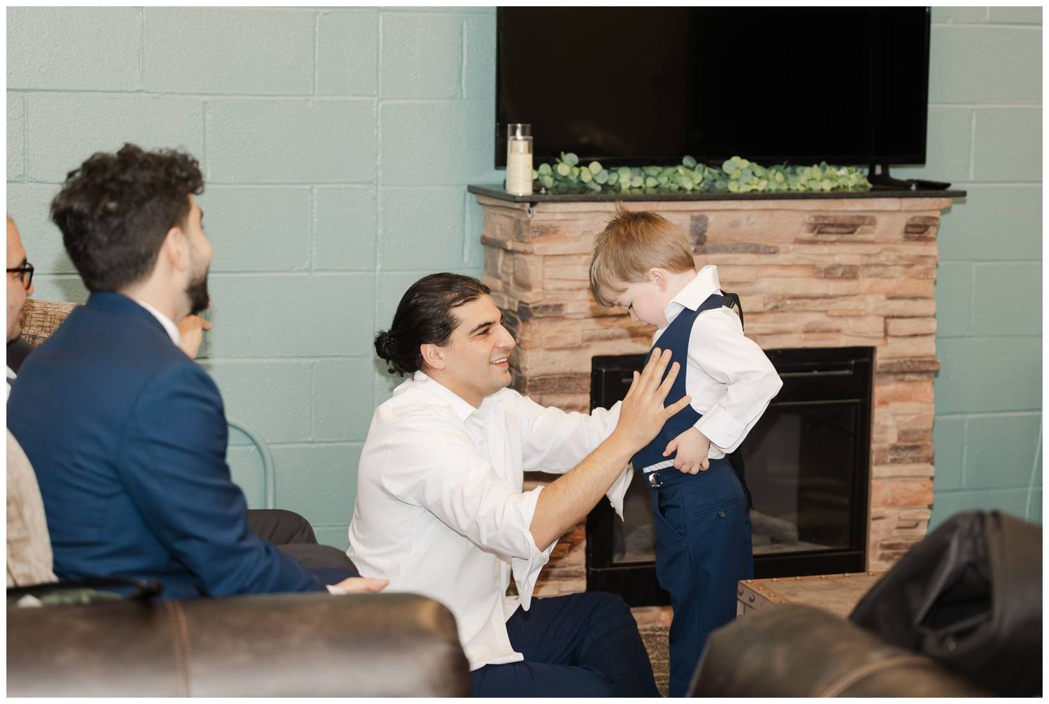 A young child in formal attire is being helped with clothing adjustments in a living room with a fireplace.