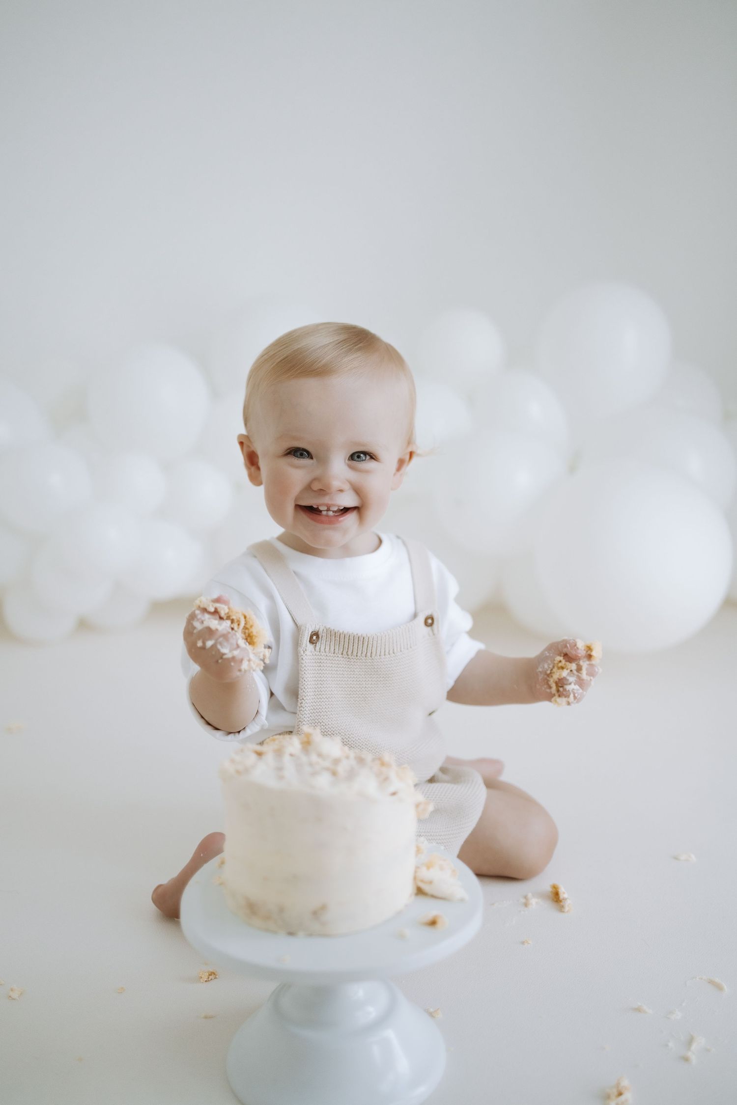 Happy baby in white outfit smiles while sitting next to white birthday cake on pedestal with balloon backdrop.