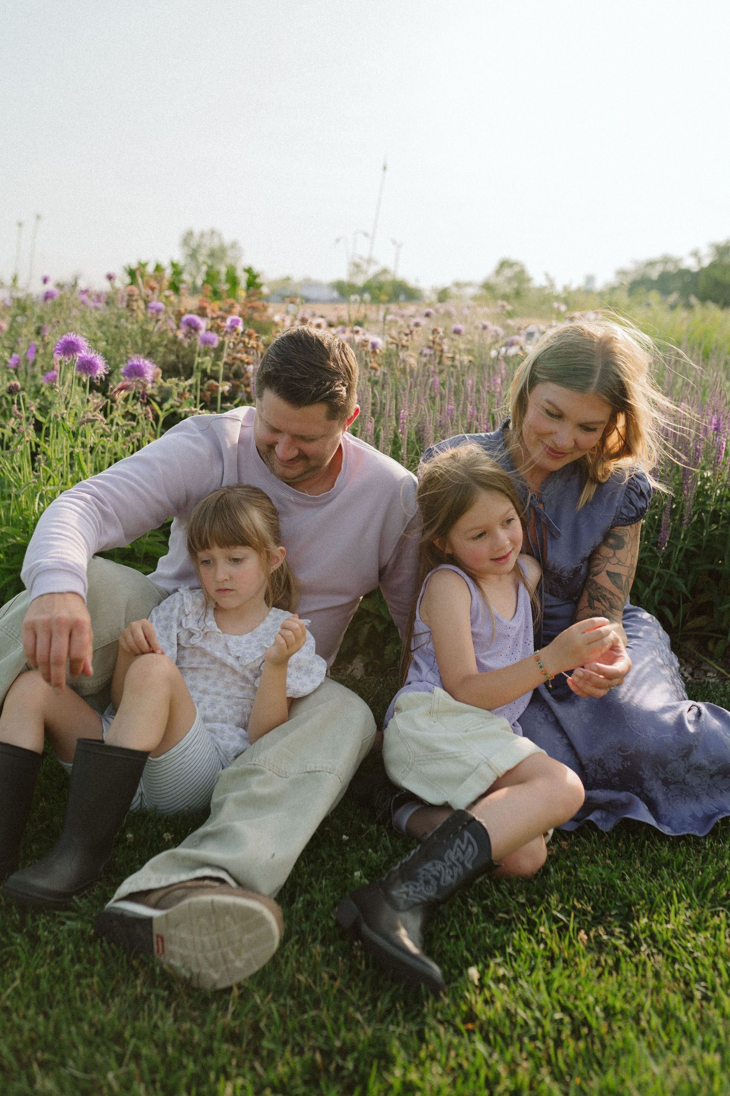 A family relaxes together on grass near blooming purple wildflowers during a sunny summer evening.