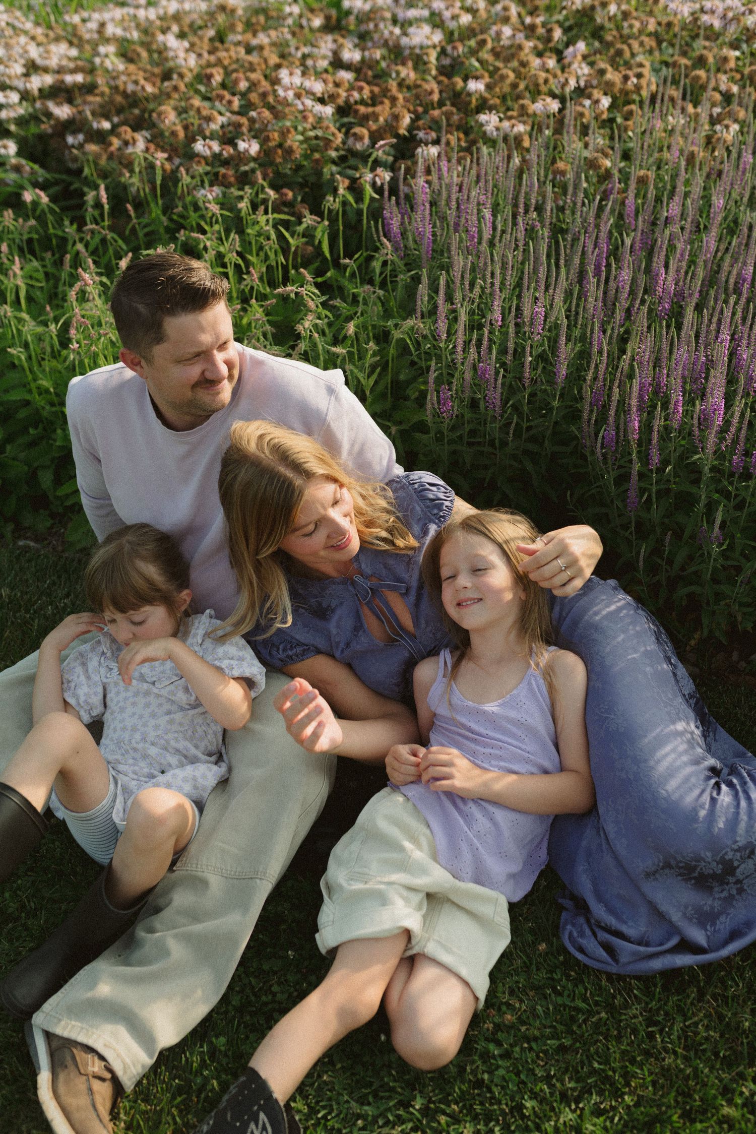 A group relaxes together on green grass with purple flowers blooming in the background on a sunny day.