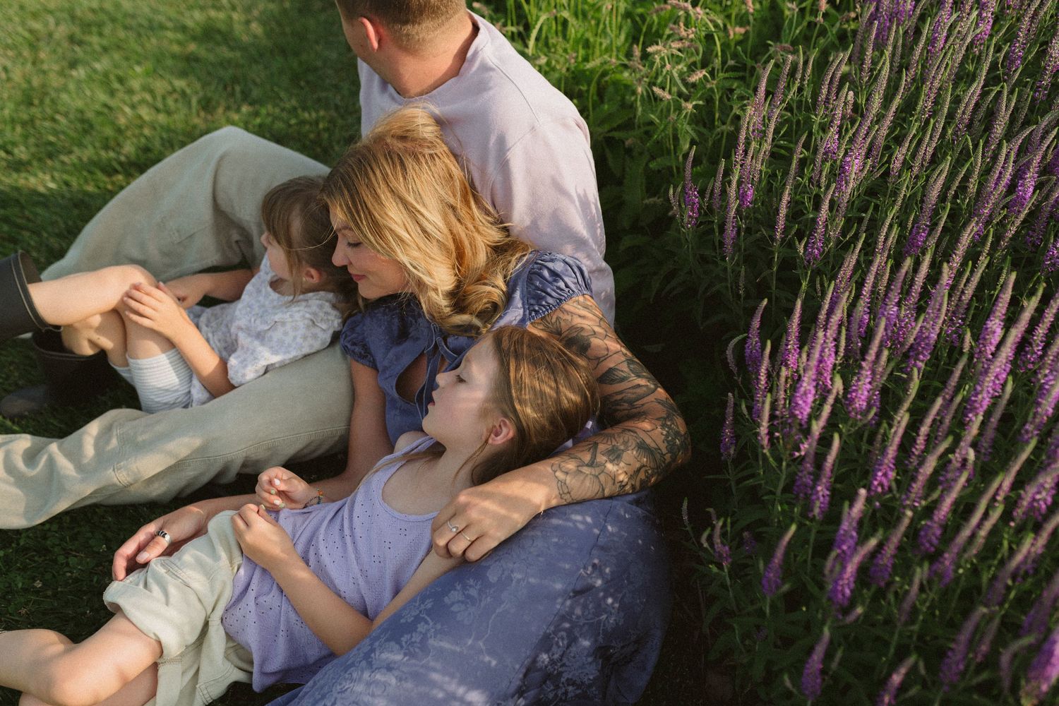 Family playfully rolling down a grassy hill together with purple wildflowers in the background.