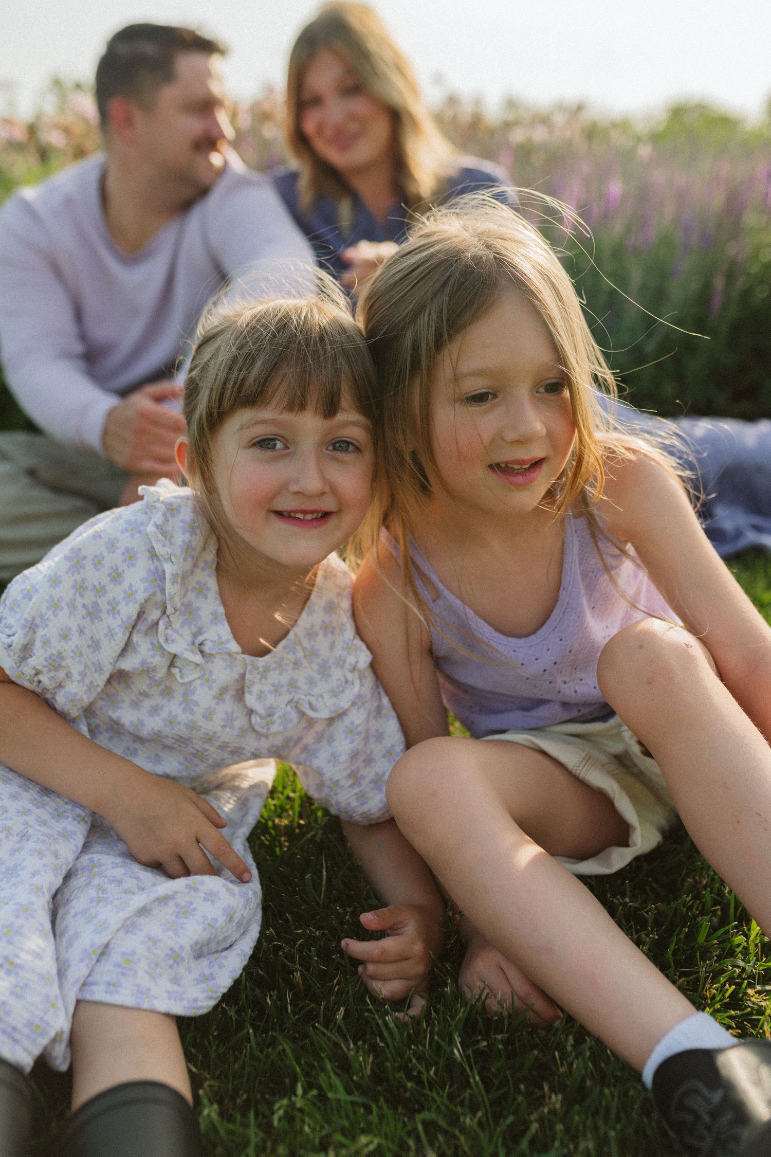 Two children sit close together on grass with purple flowers blooming in the background.