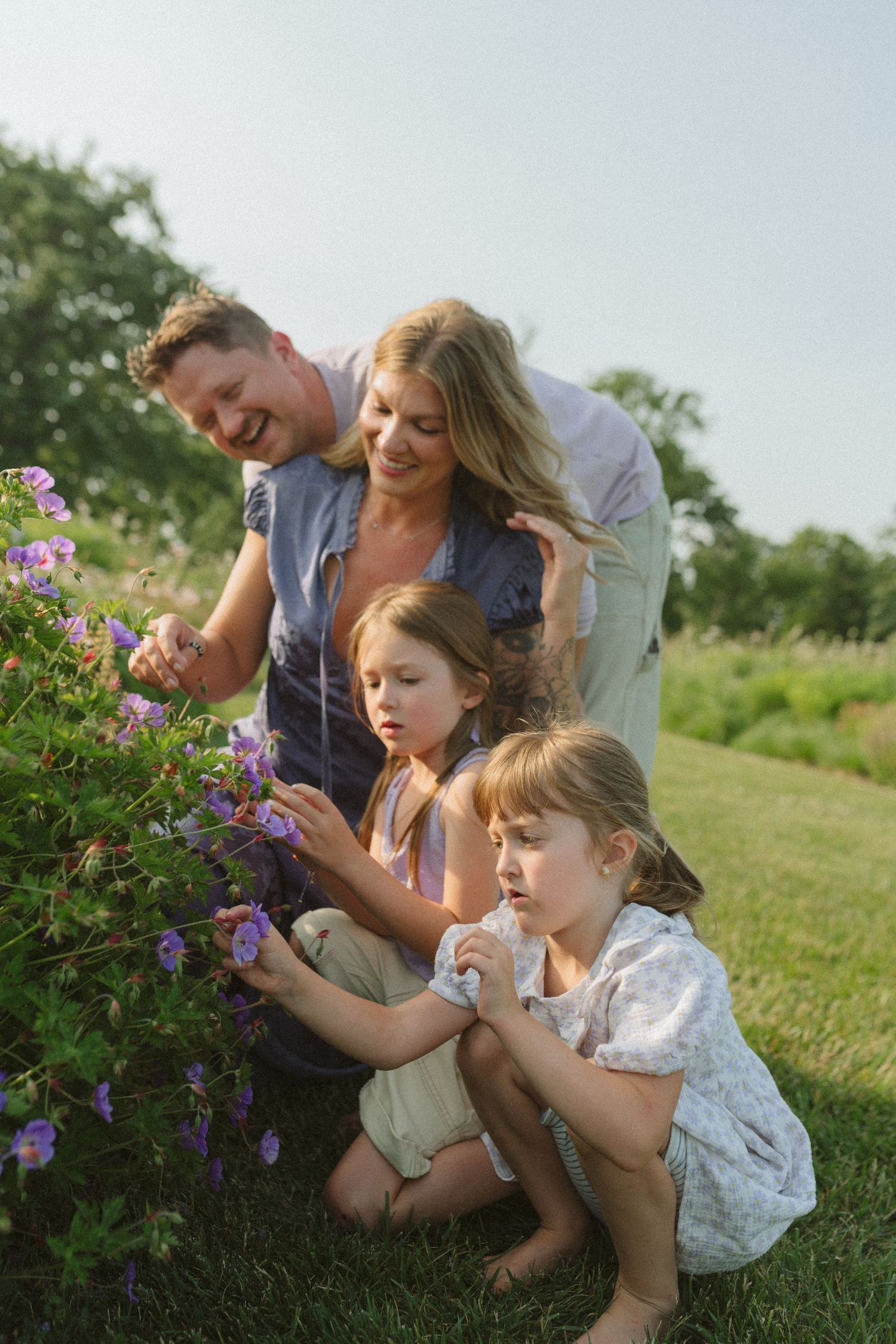 A family examines purple wildflowers together in a summer meadow.