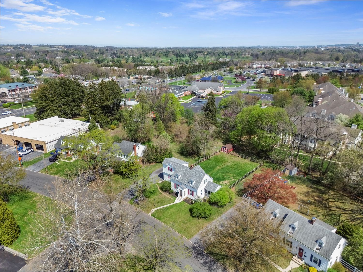 Aerial view of suburban neighborhood with green lawns, mature trees, classic homes, and commercial district visible in background.