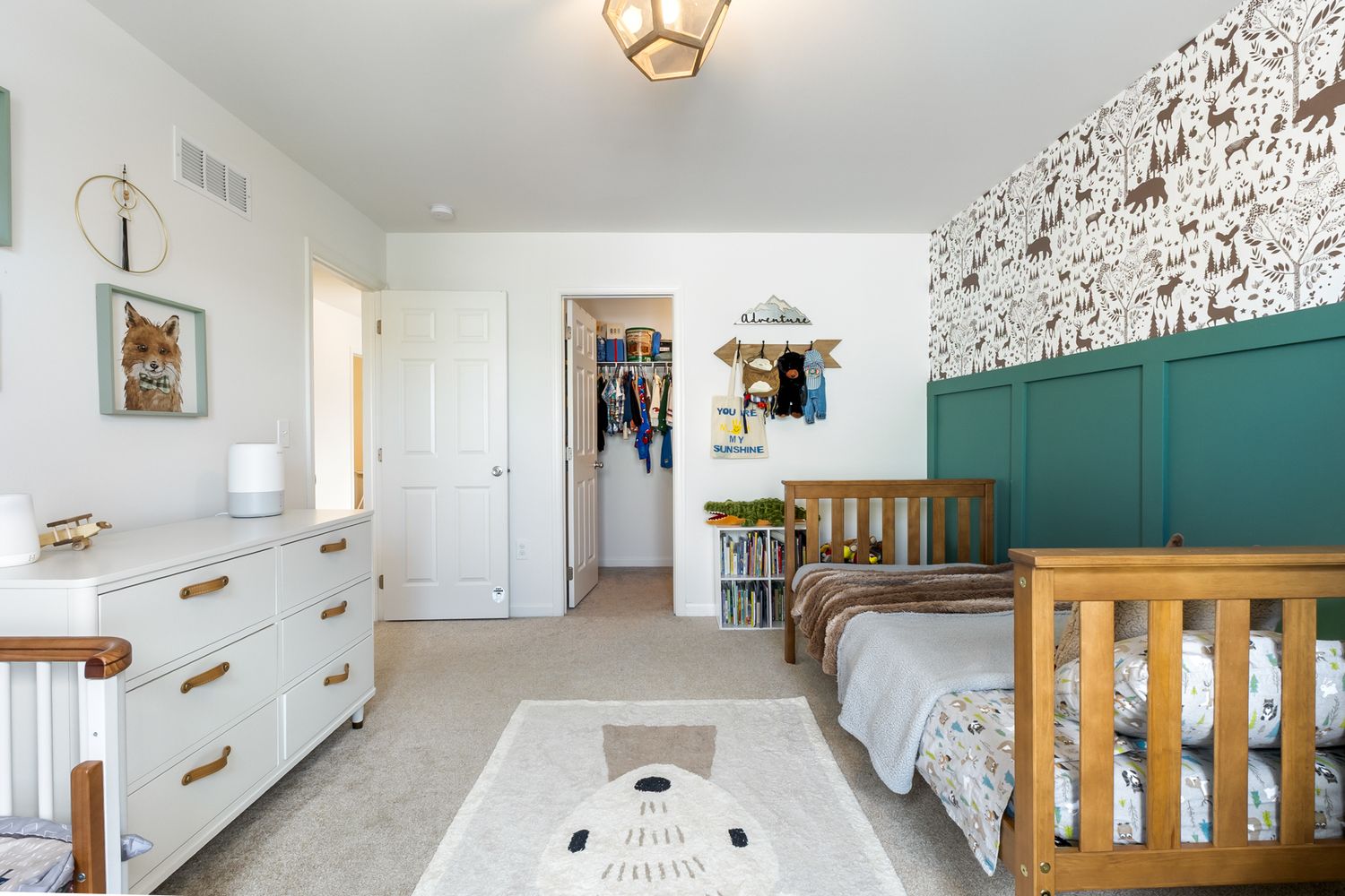 Children's bedroom with teal accent wall, wood crib, white dresser, animal wallpaper, and bear rug on neutral carpet.