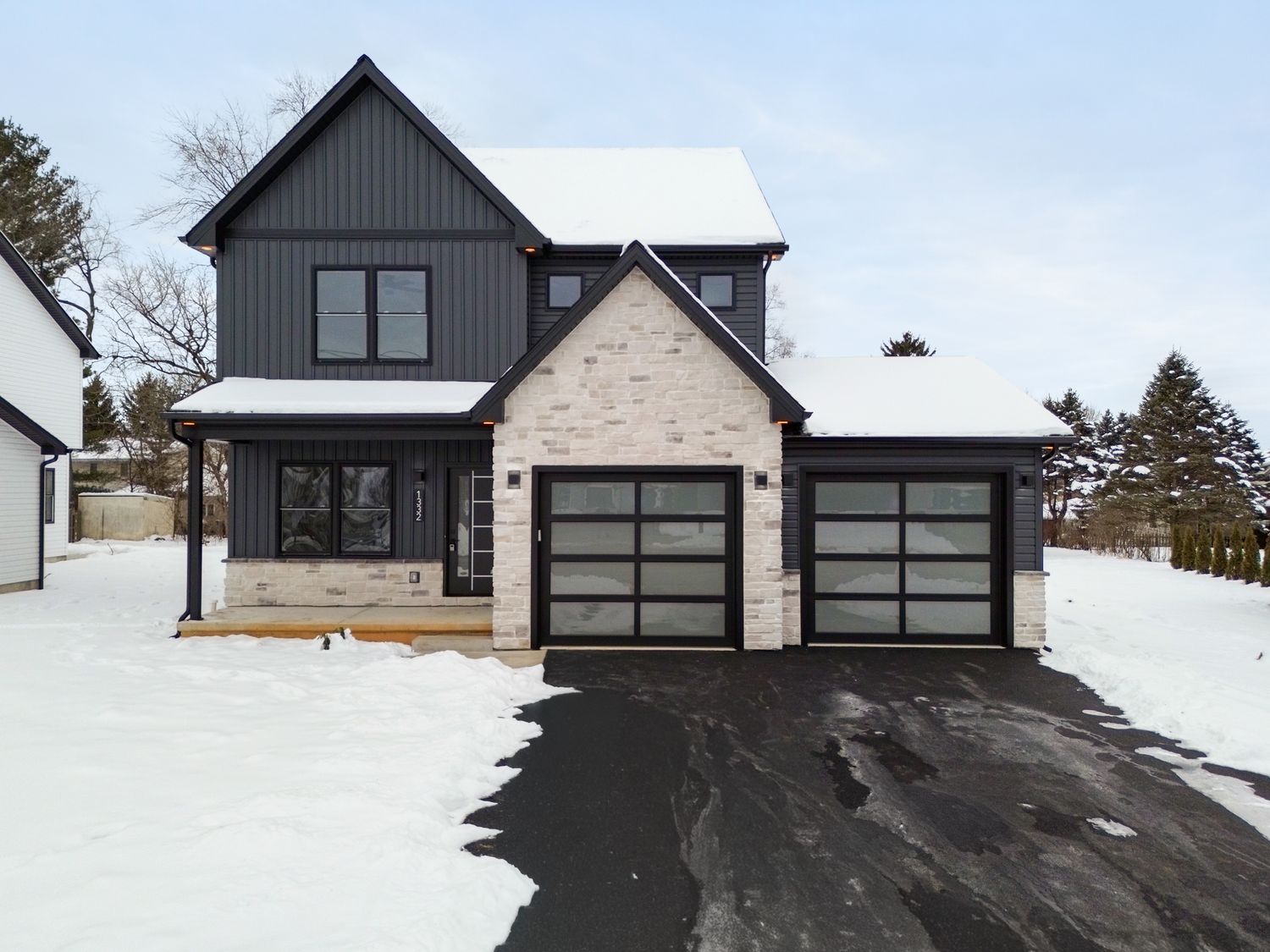 Modern black and gray farmhouse exterior with glass garage doors and snow-covered driveway in winter landscape.