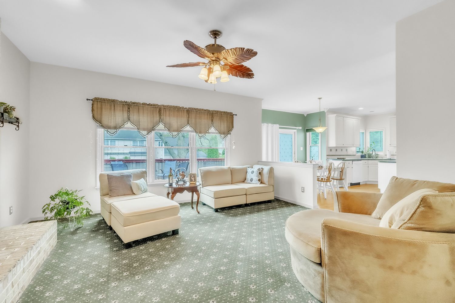 Bright living room with green patterned carpet, beige sofas, ceiling fan, and open view to kitchen.