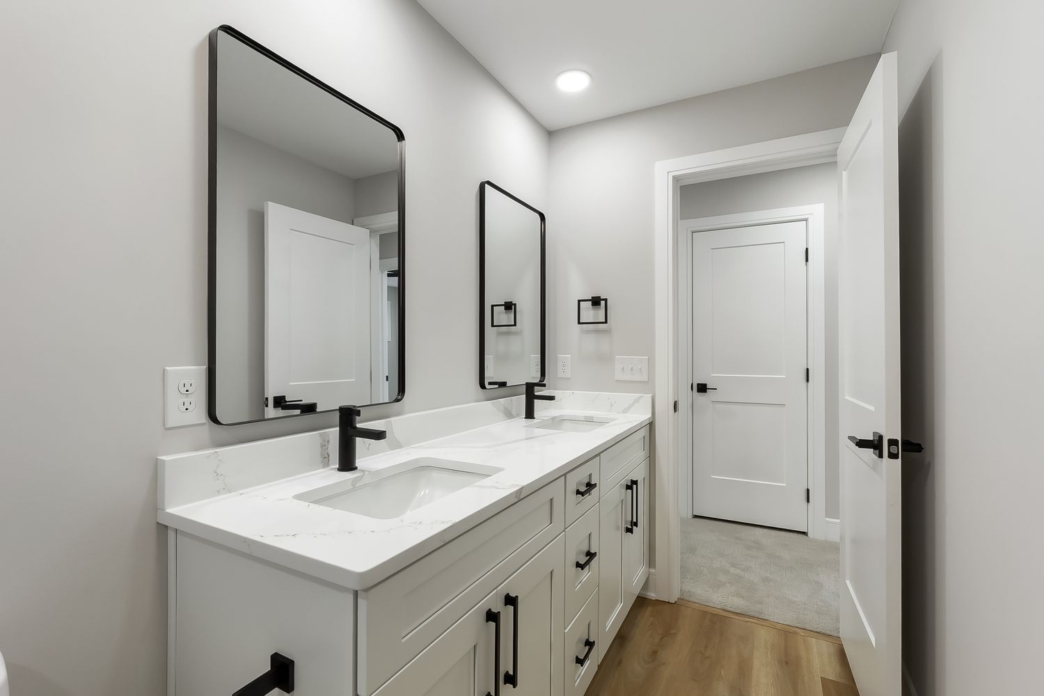 Modern white bathroom with double vanity, black hardware, hardwood floors and recessed lighting.