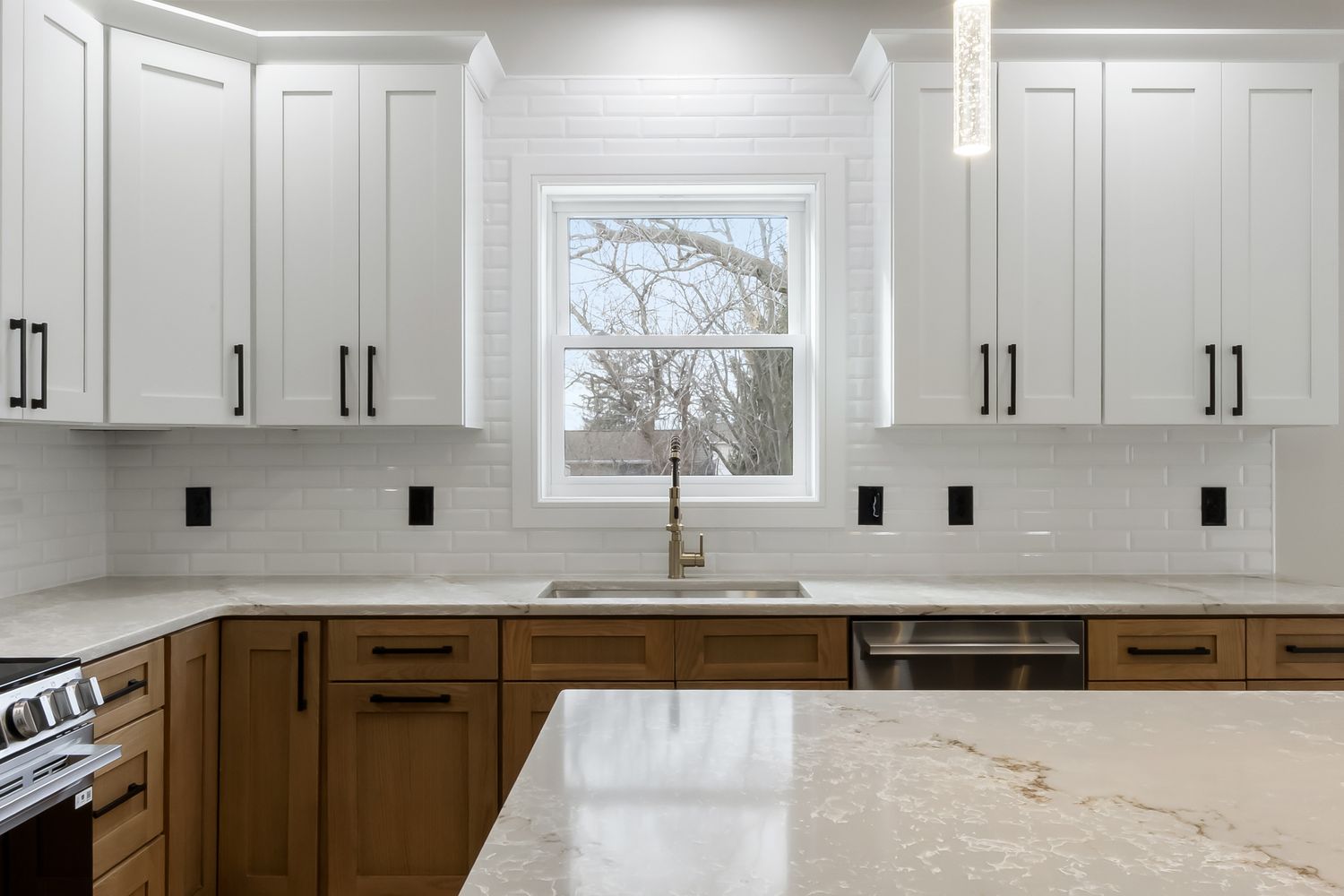 Modern kitchen with white upper cabinets, wood lower cabinets, marble countertops and a window centered above the sink.