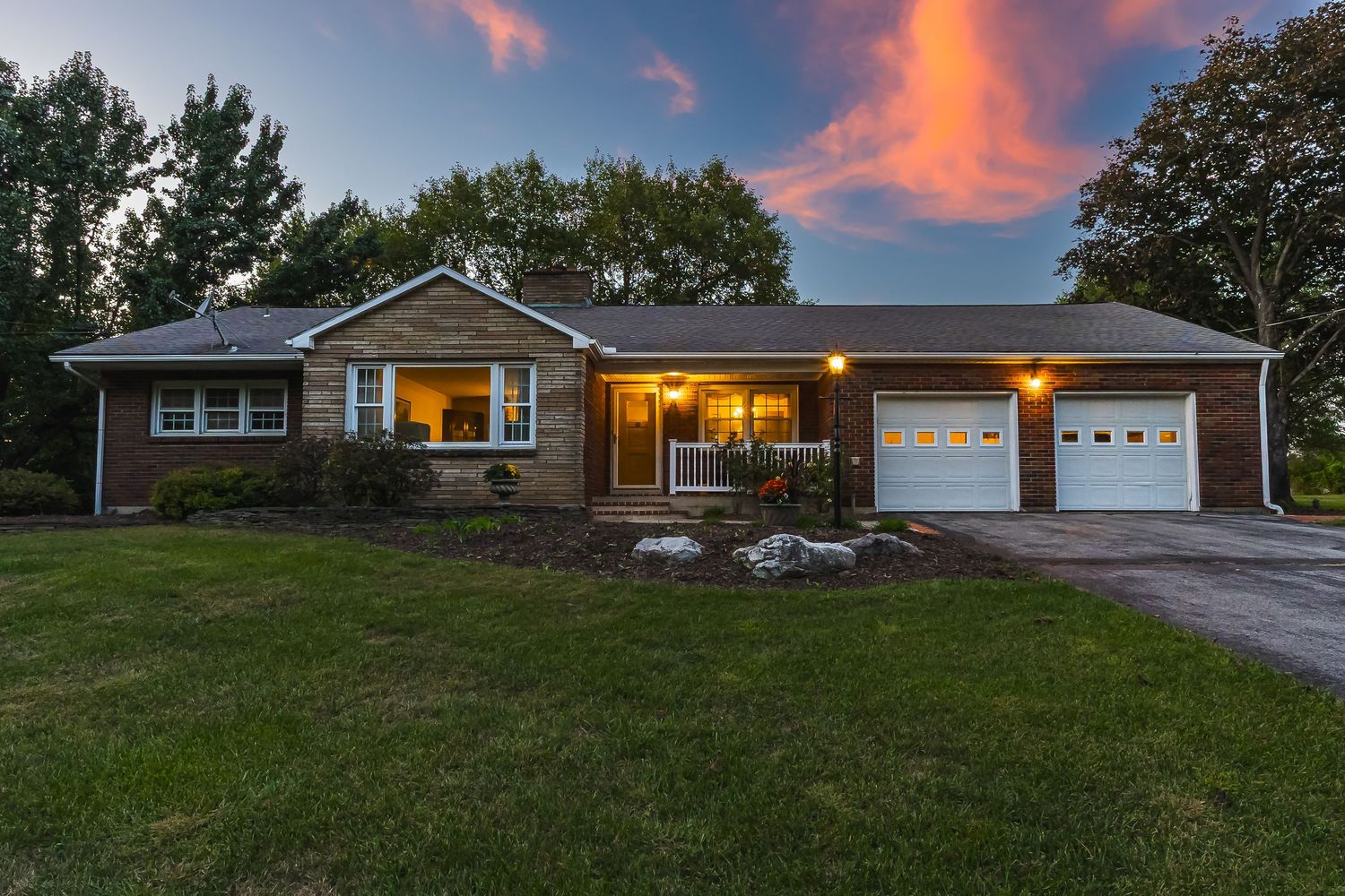 Ranch-style home with illuminated windows and attached garage at dusk with vibrant pink clouds overhead.