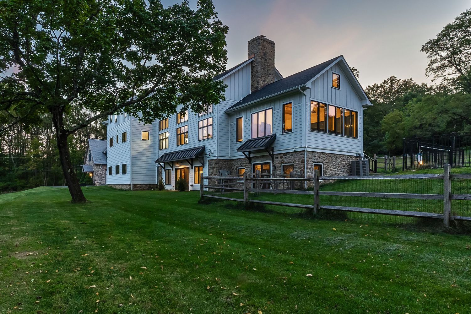 Elegant white farmhouse with stone chimney and rustic split rail fence at sunset.