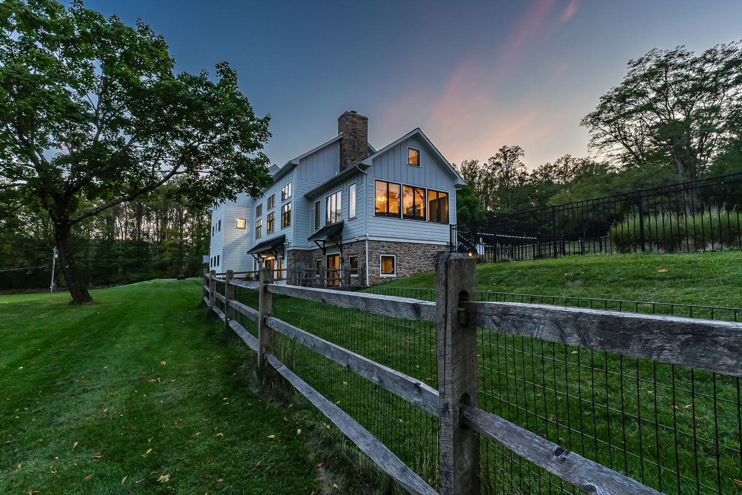 Contemporary white farmhouse with split rail fence and illuminated windows at dusk.