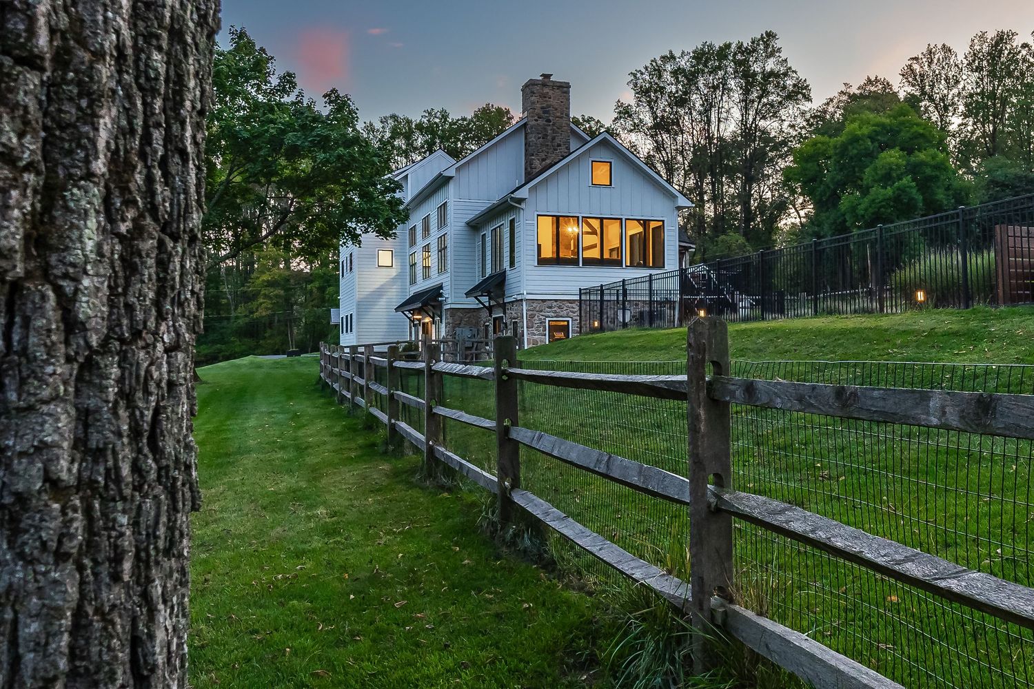 Modern farmhouse with glowing windows behind rustic wooden fence at twilight.