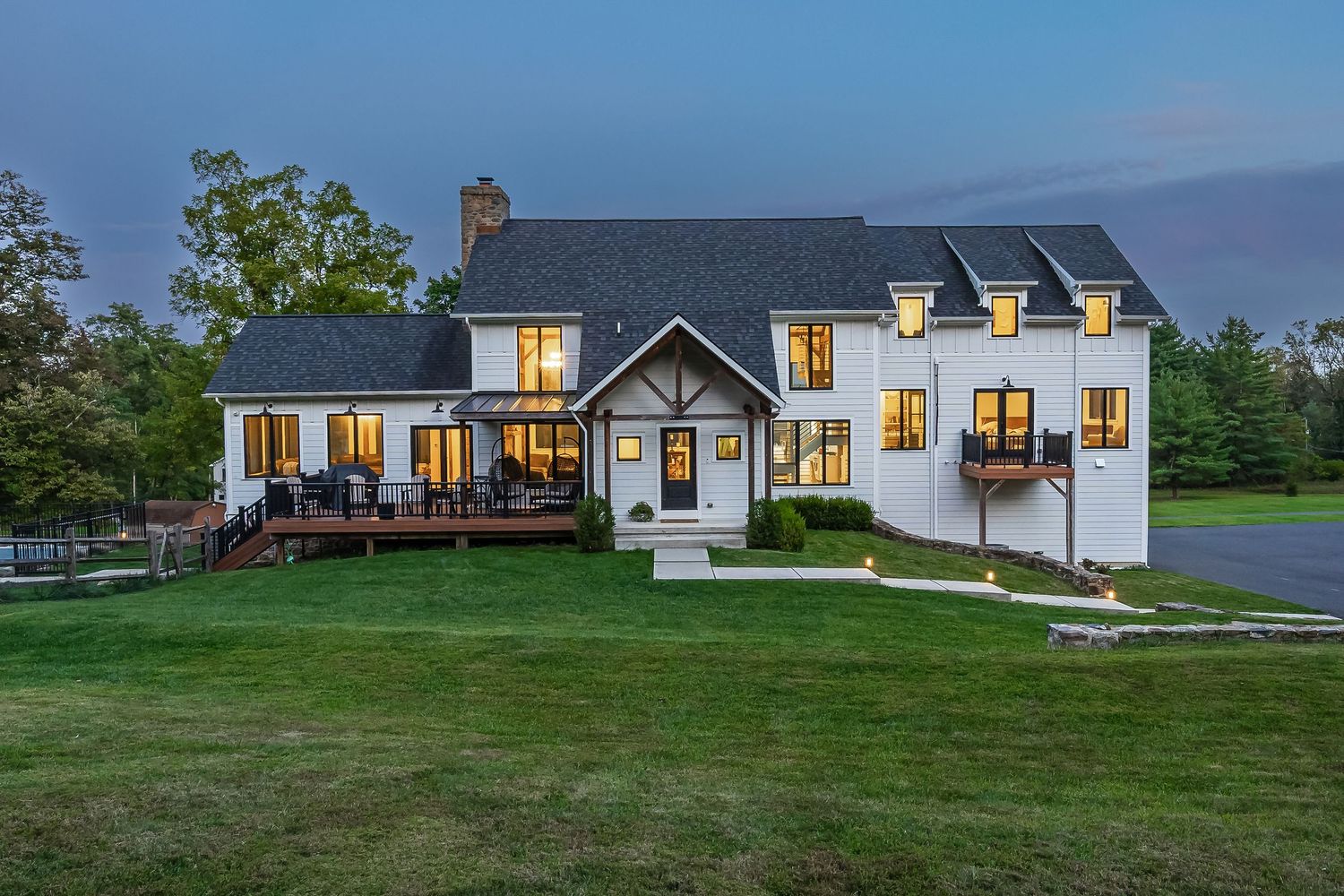 Large modern farmhouse with covered porch and multiple dormers illuminated at dusk.