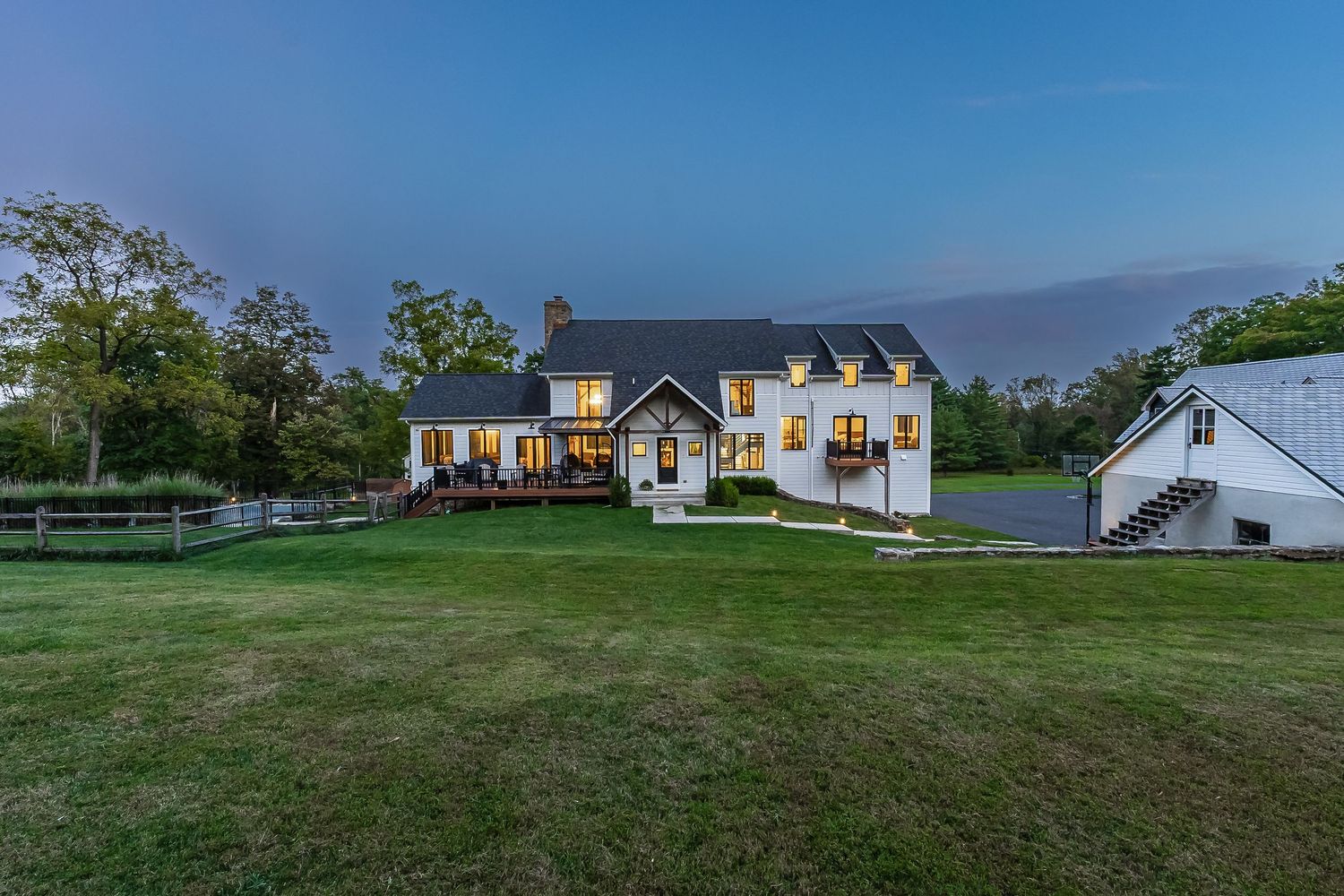 Elegant white farmhouse with attached deck and detached garage on sprawling lawn at twilight.