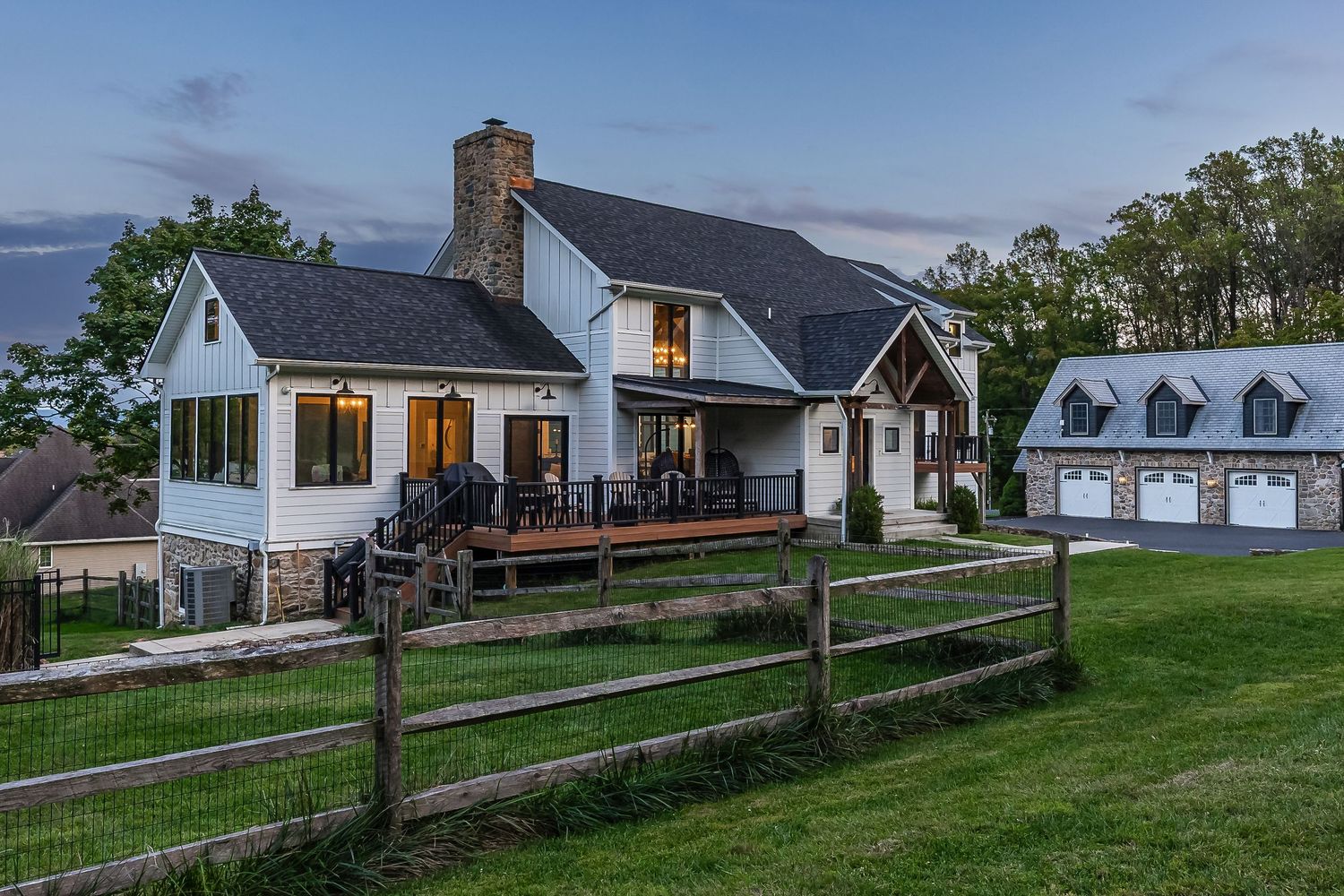 Modern white farmhouse with wraparound deck and separate garage building at dusk.