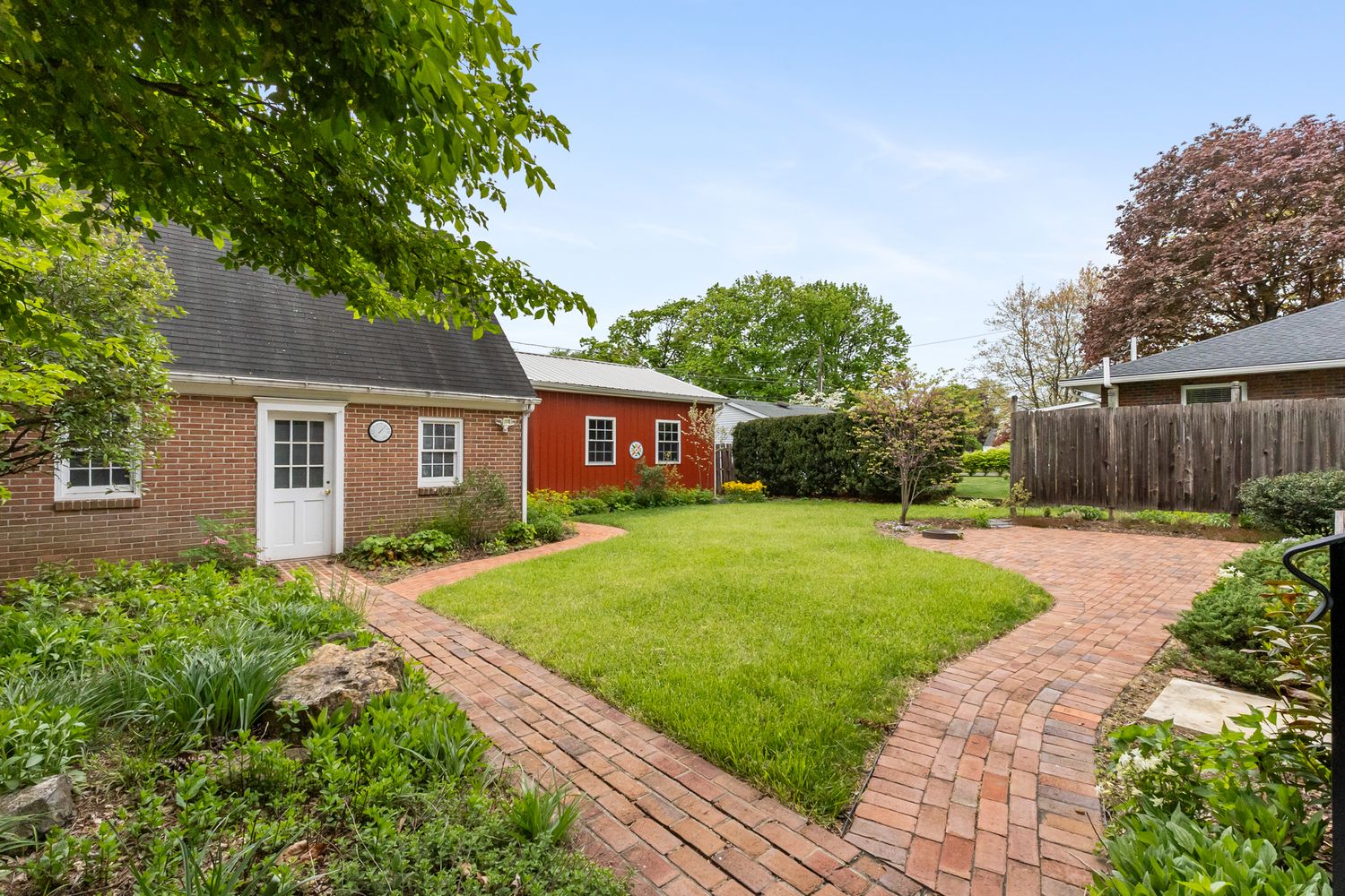 Brick pathway winds through lush green backyard with charming red outbuilding and manicured lawn beside classic brick home.