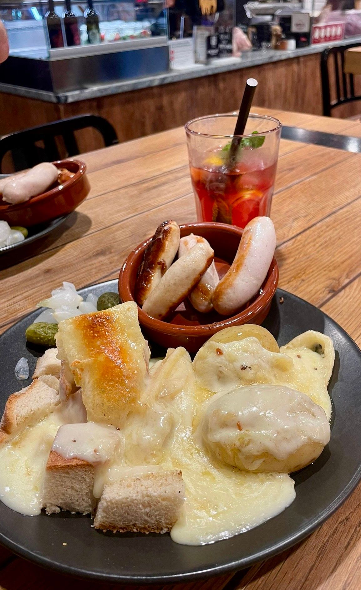 A plate of bread topped with creamy cheese sauce served with pickles and a red cocktail on a wooden table.