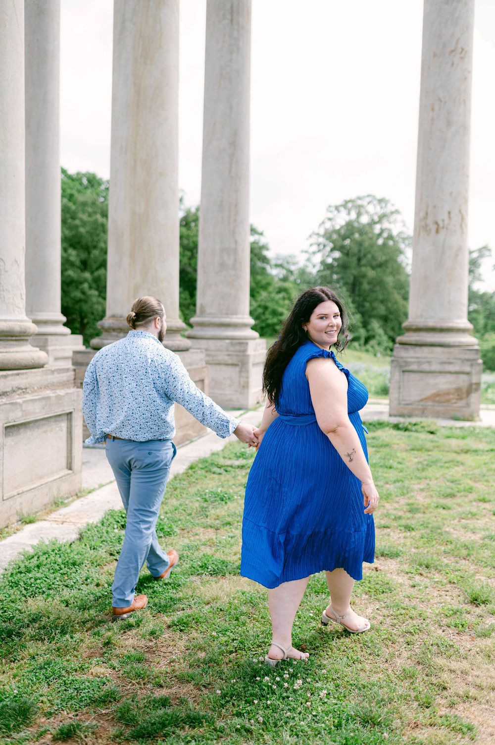 A couple holds hands while walking between white stone columns on a grassy hill in a romantic outdoor setting.