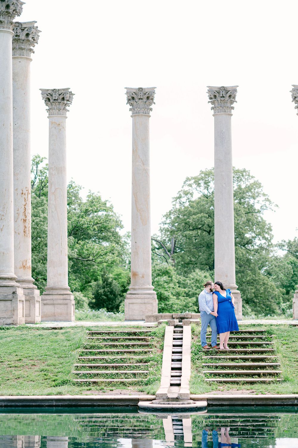 Elegant white columns and stone steps frame a reflecting pool in a romantic garden setting.