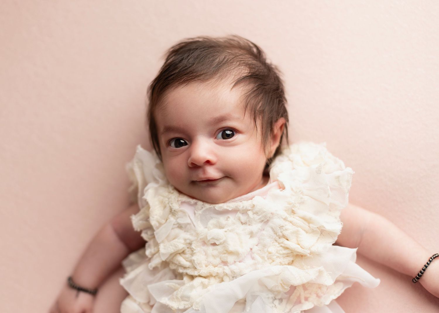 A small baby wearing a delicate cream-colored lace dress lies on a soft pink background.