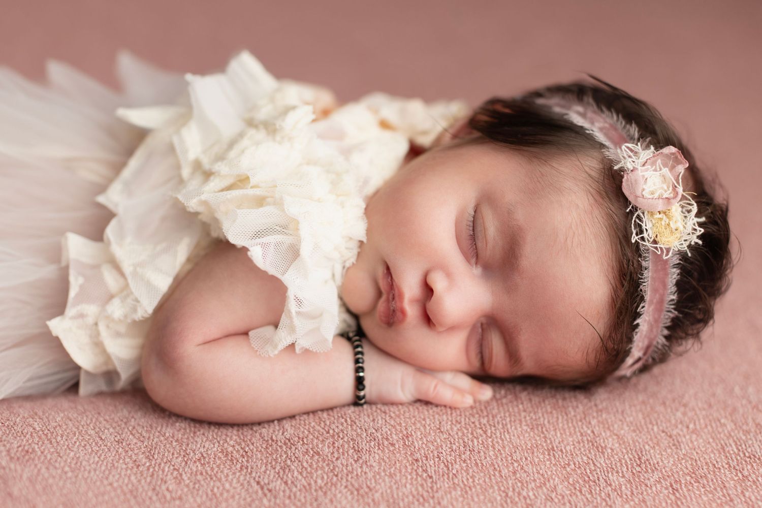 A sleeping newborn baby in a white lace outfit lies on a soft pink blanket with a delicate bow headband.