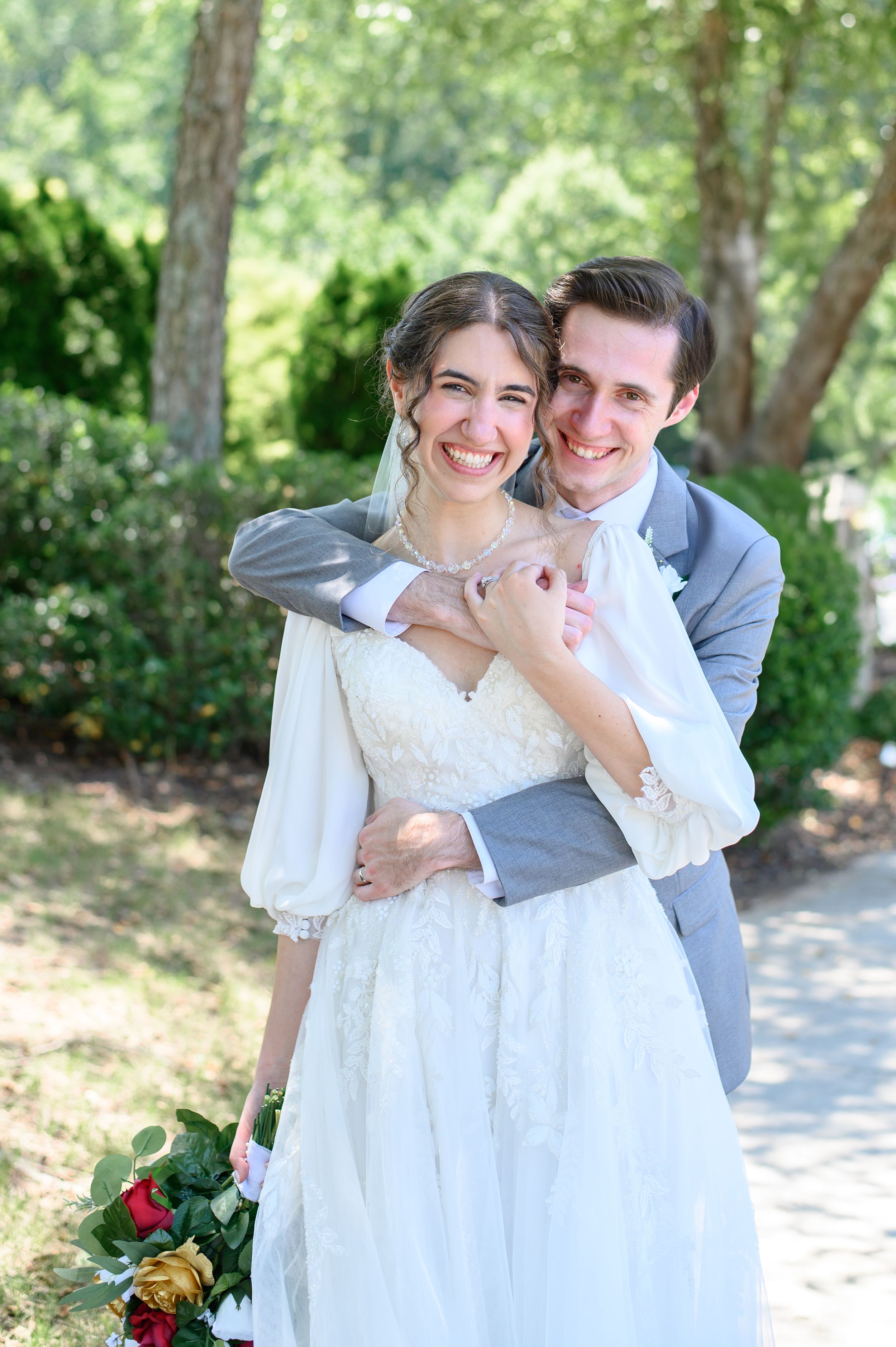 Happy bride in lace gown and groom in gray suit embracing outdoors among trees on their wedding day.