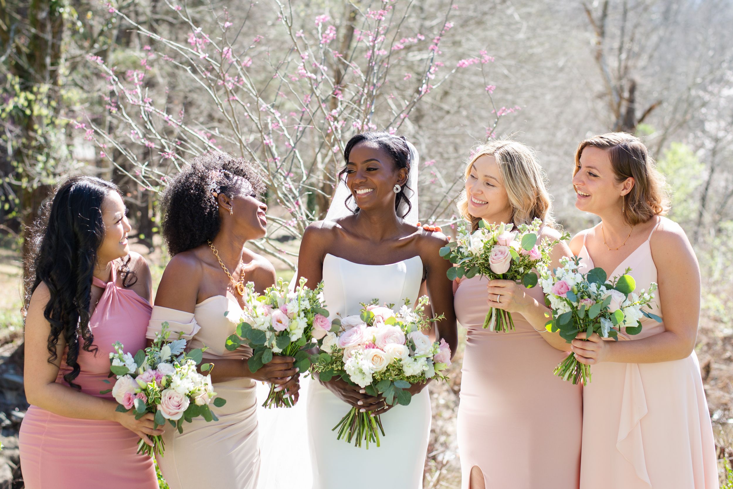 A bridal party wearing blush pink dresses holds beautiful floral bouquets while sharing a joyful moment outdoors. rays on the river wedding
