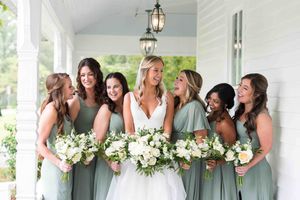 Bridesmaids in sage green dresses pose with white floral bouquets on a bright white porch.
