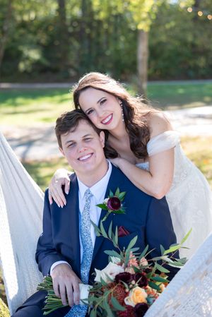 A couple in wedding attire shares intimate moments while seated on a white bench in a sunlit garden setting.