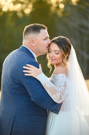 A tender moment between newlyweds at sunset, with the bride in a lace gown and flowing veil, and groom in navy suit.