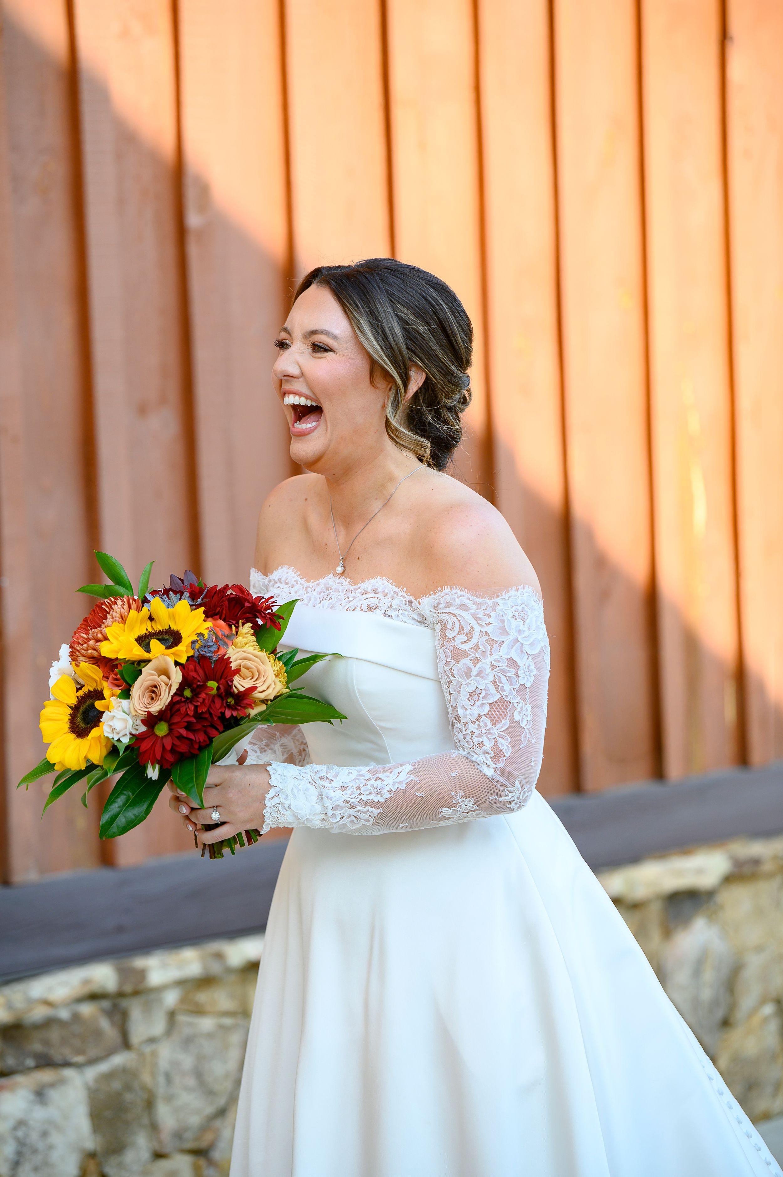 A joyful bride in a lace-sleeved white gown laughs while holding a vibrant bouquet of sunflowers and red flowers.